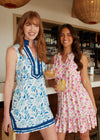 Smiling models holding a glass of champagne in the Cabana Life Bubbly Sleeveless Tunic Cover Up. A sleeveless mock neck shift dress with blue flowers on a white background and royal blue trim around the neckline and bottom hem