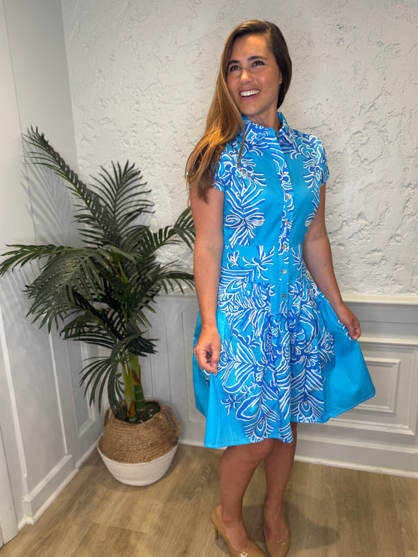 Smiling model in front of a white wall with a plant in the corner, in the Patty Kim Colette Dress - Aqua Rococo. A short sleeved mini dress with a drop waist and buttons down the front. A white and royal blue floral pattern on an aqua background