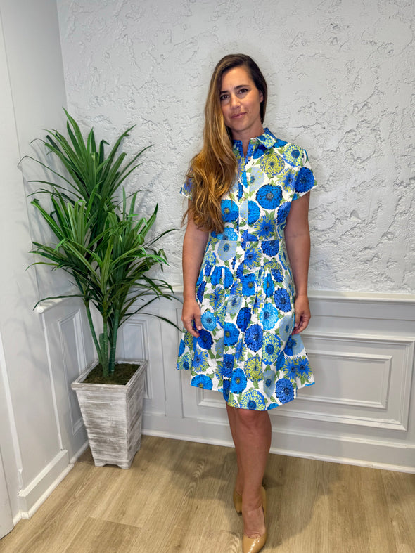 Model in front of a white wall in the Patty Kim Collette Dress - Blue Zinnias. A knee length dress with short sleeves and a drop waist and buttons down the front
