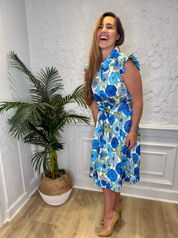 Smiling model in front of a white wall with a plant in a corner wearing the Patty Kim Megan Dress - Blue Zinnias. A midi length flutter sleeve and collar, with a tie waist and a ruffle at the bottom hem, with blue and green flower pattern