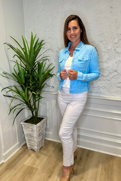 Smiling model in front of a white wall with a green plant in the corner. She's wearing the Lulu B Linen Travel Jacket - Clear Blue, a light linen jacket with 2 front pockets and lightly frayed hems. Paired with a white shirt and white pants. 