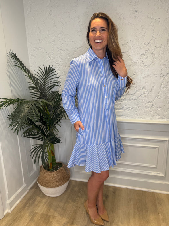 Smiling model in front of a white wall and a tropical plant in the corner wearing the Duffield Lane Iris Drop Waist Shirt Dress - Light Blue Double Stripe. A mini/knee length, long sleeve, seersucker dress. It has a collar and buttons down the front and a ruffle at the bottom hem. 