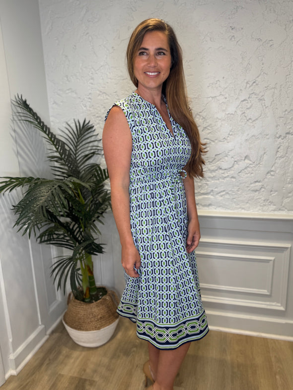 Smiling model in front of a white wall with a plant in the corner in the Jude Connally Tess Midi Dress - Terrace Geo Border Navy. A midi, v neck, sleeveless, drawstring waist dress with a white, green, and navy geometric pattern