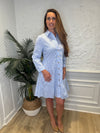 Smiling model in front of a white wall wearing the Patty Kim Capri Dress - Blue/White Stripe. A long sleeve, knee length dress that is tiered and has gold buttons down the front, with a collar. 
