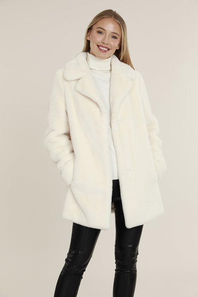 Smiling model in the Dolce Cabo Faux Fur Coat - Ivory, a faux fur mid-thigh coat, featuring a fold over collar and pockets