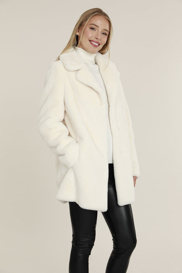 Smiling model in the Dolce Cabo Faux Fur Coat - Ivory, a faux fur mid-thigh coat, featuring a fold over collar and pockets