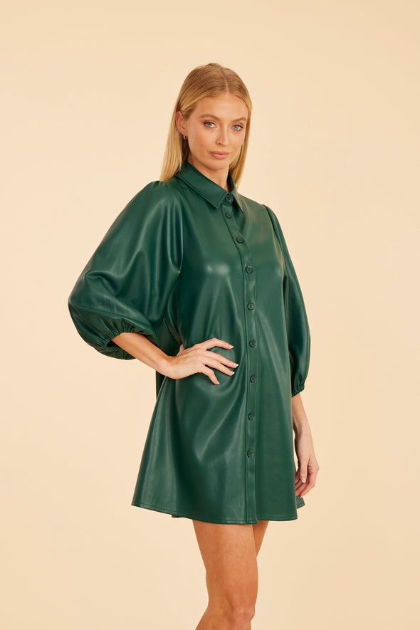 Model with hand on hip is wearing the Dolce Cabo Soft Vegan Leather Dress in Forest Green. A mini shift silhouette with soft faux leather, 3/4 puff sleeves, and front button detailing