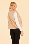 Smiling model in the Dolce Cabo Faux Fur Vest - Champagne. A light beige fur vest with a fold over collar and pockets over a white long-sleeve shirt and black pants on a beige background