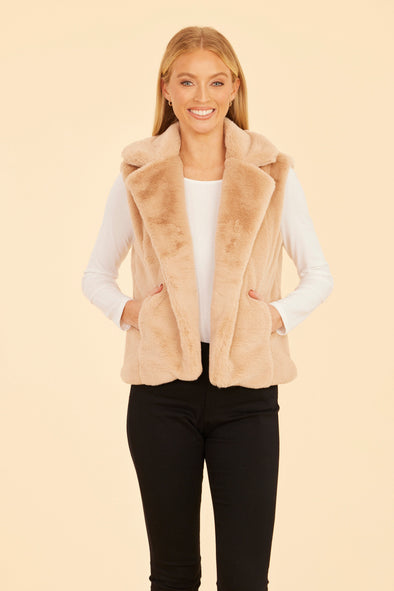 Smiling model in the Dolce Cabo Faux Fur Vest - Champagne. A light beige fur vest with a fold over collar and pockets. 