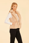 Smiling model with hands in pockets in the Dolce Cabo Faux Fur Vest - Champagne. A light beige fur vest with a fold over collar and pockets. 