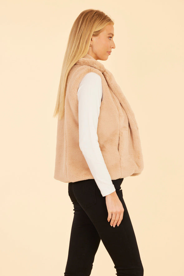 Side view of model in the Dolce Cabo Faux Fur Vest - Champagne. A light beige fur vest with a fold over collar and pockets. 
