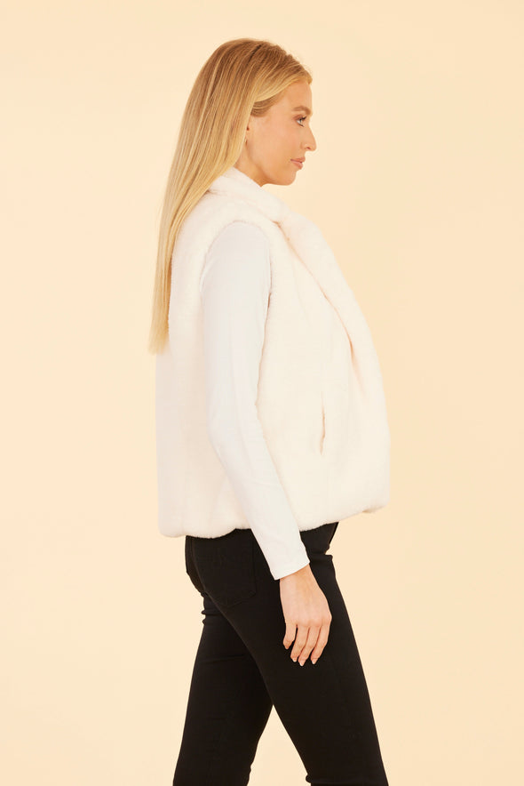 Smiling model with hands in her pockets in the Dolce Cabo Faux Fur Vest - White, a white faux fur vest with a fold over collar and pockets 