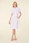 Smiling model in a knee length pink seersucker dress with a collar, pockets on the chest, and a tie belt