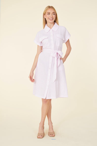 Smiling model in a knee length pink seersucker dress with a collar, pockets on the chest, and a tie belt