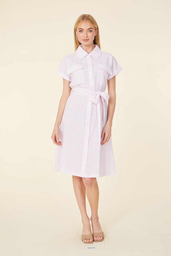 Smiling model in a knee length pink seersucker dress with a collar, pockets on the chest, and a tie belt 