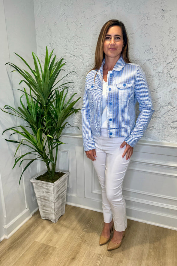 Smiling model in front of a white wall with a green plant in the corner. She's wearing the Lulu B Linen Travel Jacket - Seersucker, a light linen jacket with 2 front pockets and lightly frayed hems. Paired with a white shirt and white pants. 