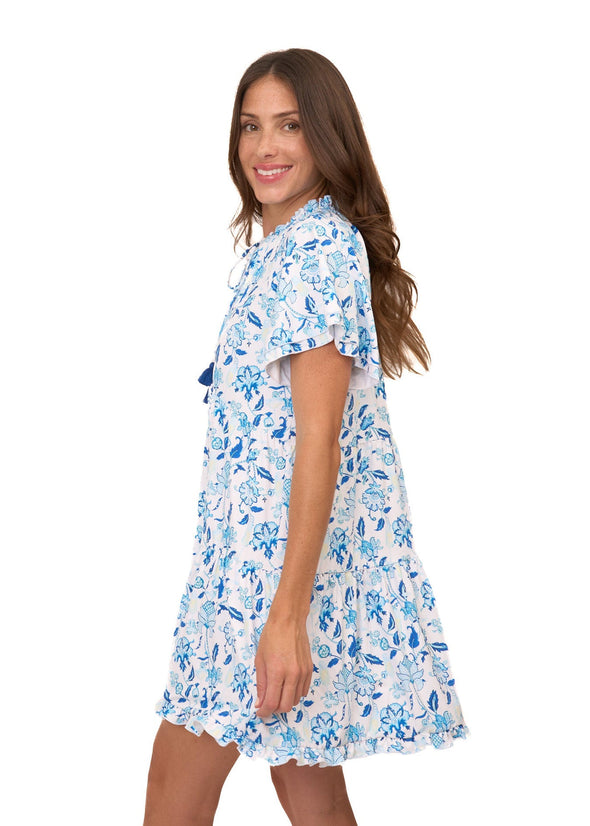 Smiling model holding a glass of champagne in the Cabana Life Bubbly Short Sleeve Tiered Ruffle Dress. A short sleeved, tiered, v-neck dress that is white with blue floral pattern 