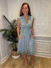 Smiling model in front of a white wall with a plant in the corner in the Jude Connally Tess Midi Dress - Terrace Geo Border Navy. A midi, v neck, sleeveless, drawstring waist dress with a white, green, and navy geometric pattern
