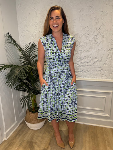 Smiling model in front of a white wall with a plant in the corner in the Jude Connally Tess Midi Dress - Terrace Geo Border Navy. A midi, v neck, sleeveless, drawstring waist dress with a white, green, and navy geometric pattern
