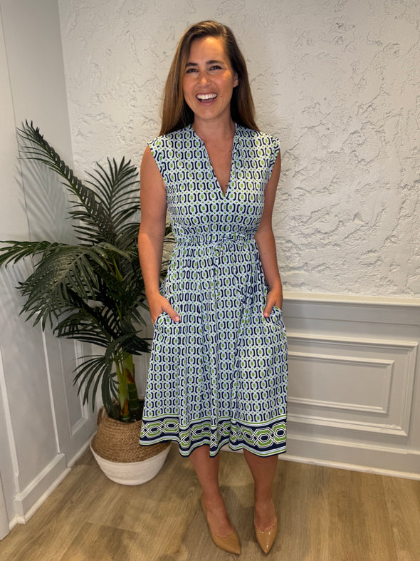 Smiling model in front of a white wall with a plant in the corner in the Jude Connally Tess Midi Dress - Terrace Geo Border Navy. A midi, v neck, sleeveless, drawstring waist dress with a white, green, and navy geometric pattern