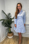 Model in front of a white wall wearing the Patty Kim Capri Dress - Blue/White Stripe. A long sleeve, knee length dress that is tiered and has gold buttons down the front, with a collar. 