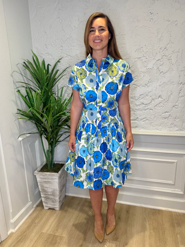 Model in front of a white wall in the Patty Kim Collette Dress - Blue Zinnias. A knee length dress with short sleeves and a drop waist and buttons down the front