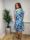 Back view of the model in front of a white wall in the Patty Kim Collette Dress - Blue Zinnias. A knee length dress with short sleeves and a drop waist and buttons down the front