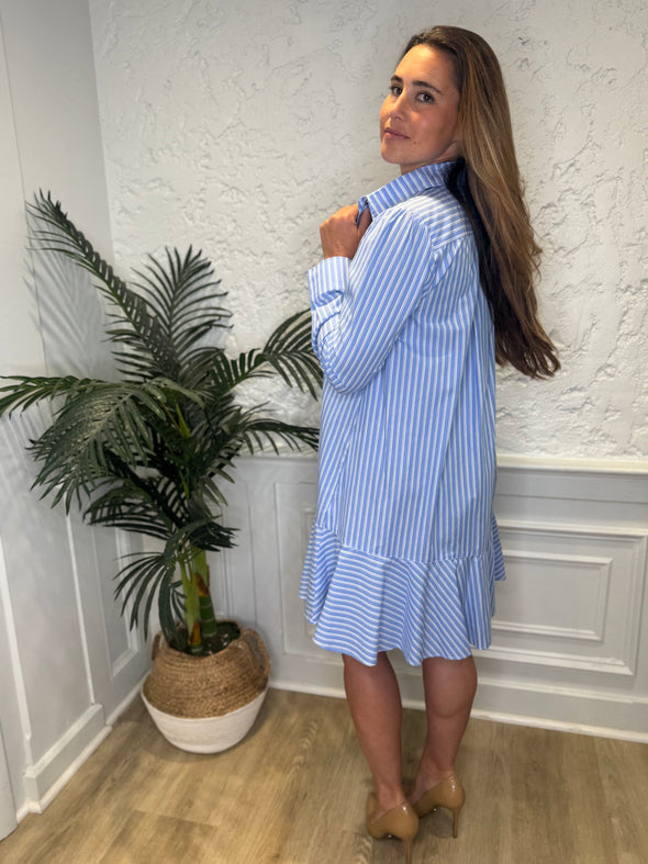 Smiling model in front of a white wall and a tropical plant in the corner wearing the Duffield Lane Iris Drop Waist Shirt Dress - Light Blue Double Stripe. A mini/knee length, long sleeve, seersucker dress. It has a collar and buttons down the front and a ruffle at the bottom hem. 