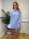 Smiling model twirling in front of a white wall and a tropical plant in the corner wearing the Duffield Lane Iris Drop Waist Shirt Dress - Light Blue Double Stripe. A mini/knee length, long sleeve, seersucker dress. It has a collar and buttons down the front and a ruffle at the bottom hem. 