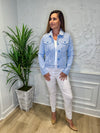 Smiling model in front of a white wall with a green plant in the corner. She's wearing the Lulu B Linen Travel Jacket - Seersucker, a light linen jacket with 2 front pockets and lightly frayed hems. Paired with a white shirt and white pants. 