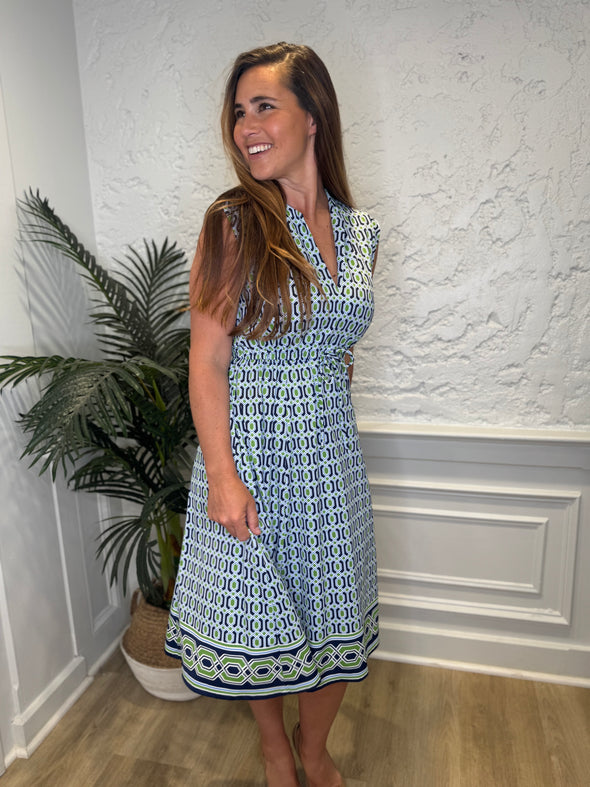 Smiling model in front of a white wall with a plant in the corner in the Jude Connally Tess Midi Dress - Terrace Geo Border Navy. A midi, v neck, sleeveless, drawstring waist dress with a white, green, and navy geometric pattern