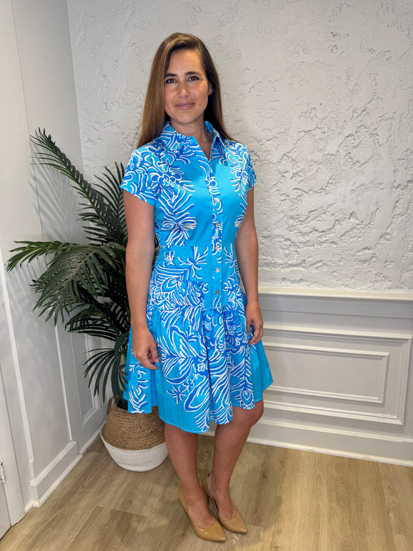 Smiling model in front of a white wall with a plant in the corner, in the Patty Kim Colette Dress - Aqua Rococo. A short sleeved mini dress with a drop waist and buttons down the front. A white and royal blue floral pattern on an aqua background