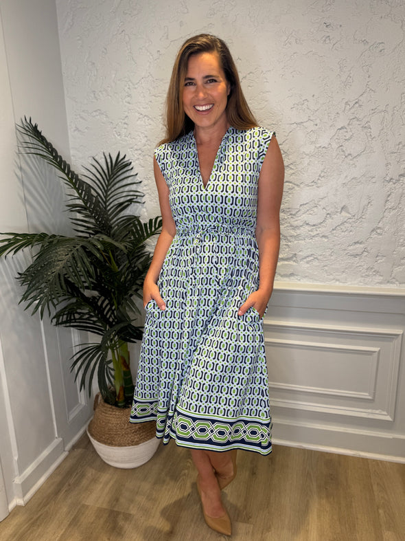 Smiling model in front of a white wall with a plant in the corner in the Jude Connally Tess Midi Dress - Terrace Geo Border Navy. A midi, v neck, sleeveless, drawstring waist dress with a white, green, and navy geometric pattern