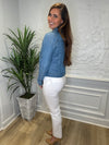 Side view of smiling model in the Lulu B Denim Travel Jacket - Blue Jean in front of a white wall with a plant in the corner. A lightweight chambray jacket paired with a white shirt and white pants
