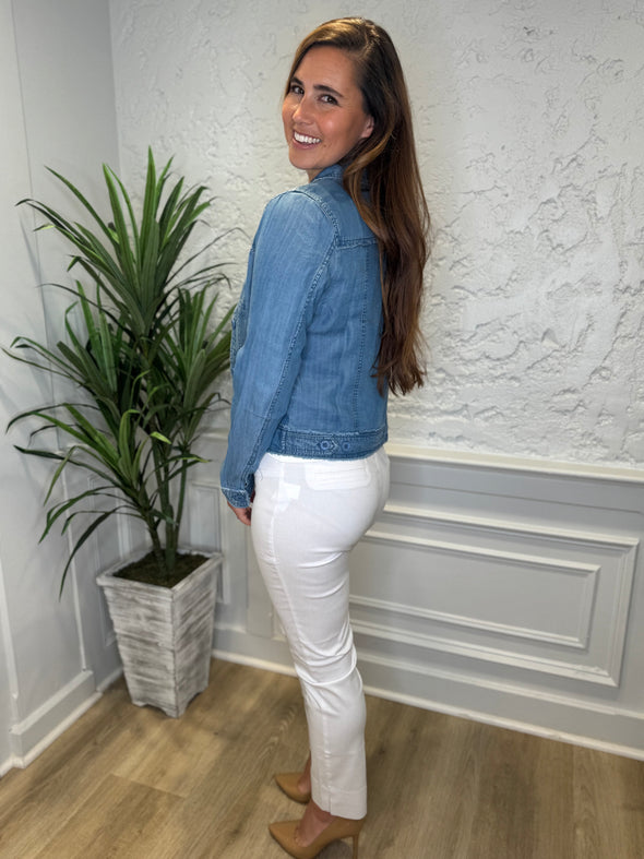Side view of smiling model in the Lulu B Denim Travel Jacket - Blue Jean in front of a white wall with a plant in the corner. A lightweight chambray jacket paired with a white shirt and white pants