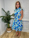 Smiling model with hands in pockets in front of a white wall with a plant in a corner wearing the Patty Kim Megan Dress - Blue Zinnias. A midi length flutter sleeve and collar, with a tie waist and a ruffle at the bottom hem, with blue and green flower pattern