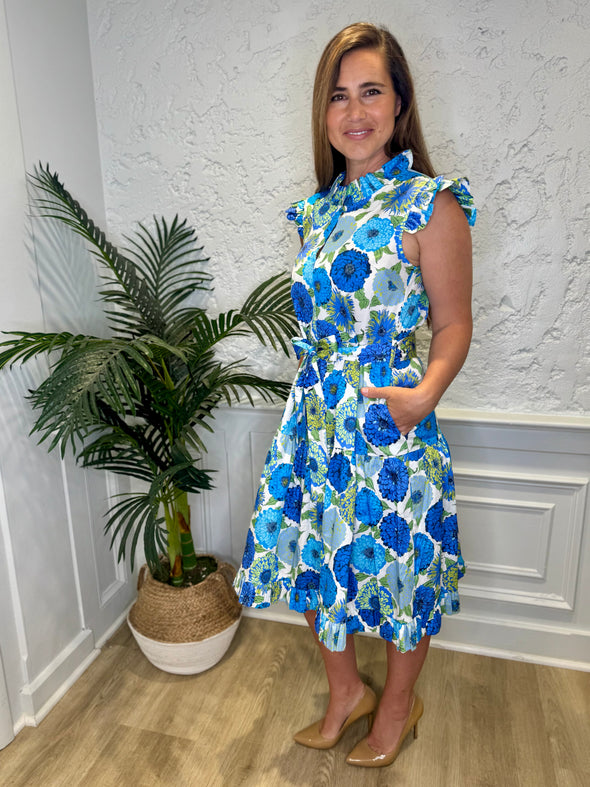 Smiling model with hands in pockets in front of a white wall with a plant in a corner wearing the Patty Kim Megan Dress - Blue Zinnias. A midi length flutter sleeve and collar, with a tie waist and a ruffle at the bottom hem, with blue and green flower pattern