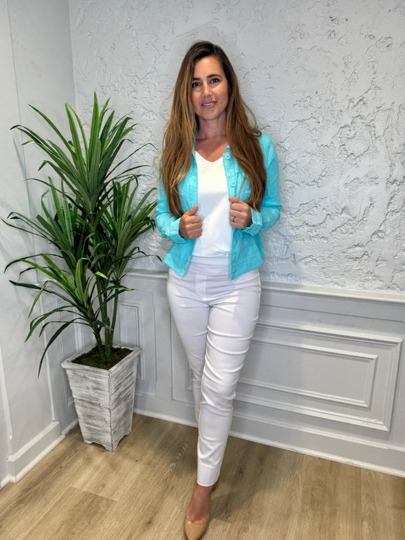 Smiling model in front of a white wall with a green plant in the corner. She's wearing the Lulu B Linen Travel Jacket - Seafoam, a light linen jacket with 2 front pockets and lightly frayed hems. Paired with a white shirt and white pants. 