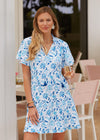 Smiling model holding a glass of champagne in the Cabana Life Bubbly Short Sleeve Tiered Ruffle Dress. A short sleeved, tiered, v-neck dress that is white with blue floral pattern 