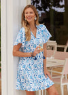 Smiling model holding a glass of champagne in the Cabana Life Bubbly Short Sleeve Tiered Ruffle Dress. A short sleeved, tiered, v-neck dress that is white with blue floral pattern 
