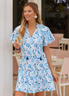 Smiling model holding a glass of champagne in the Cabana Life Bubbly Short Sleeve Tiered Ruffle Dress. A short sleeved, tiered, v-neck dress that is white with blue floral pattern 