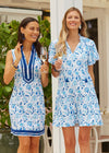Smiling models holding a glass of champagne in the Cabana Life Bubbly Sleeveless Tunic Cover Up. A sleeveless mock neck shift dress with blue flowers on a white background and royal blue trim around the neckline and bottom hem
