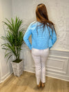 Back view of model in front of a white wall with a green plant in the corner. She's wearing the Lulu B Linen Travel Jacket - Clear Blue, a light linen jacket with 2 front pockets and lightly frayed hems. Paired with a white shirt and white pants. 