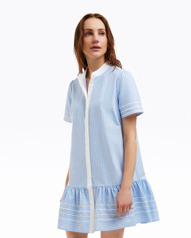 Model in the Draper James Molly Mini Dress - Oxford Shirting. A blue and white striped dress with a white collar and button cover down the front, a ruffle hem and short sleeves with ric rac detailing on hem and sleeves