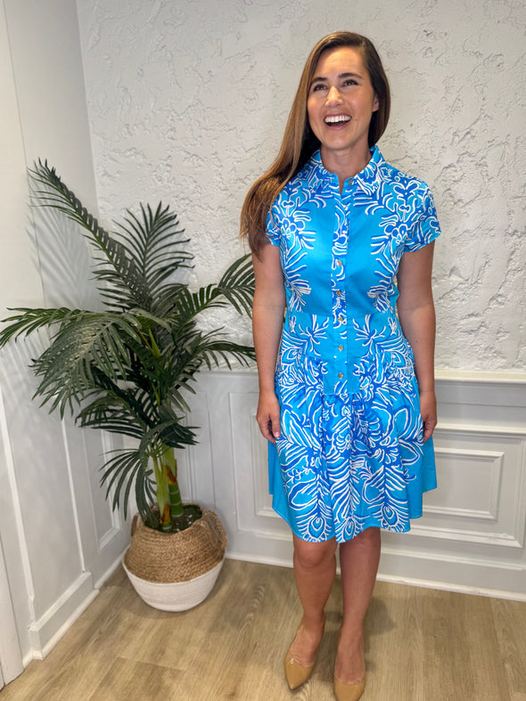 Smiling model in front of a white wall with a plant in the corner, in the Patty Kim Colette Dress - Aqua Rococo. A short sleeved mini dress with a drop waist and buttons down the front. A white and royal blue floral pattern on an aqua background