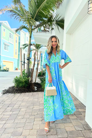 Model in a green and blue tiered maxi dress with a tropical pattern. Also has a v-neck with a traditional collar and short sleeves