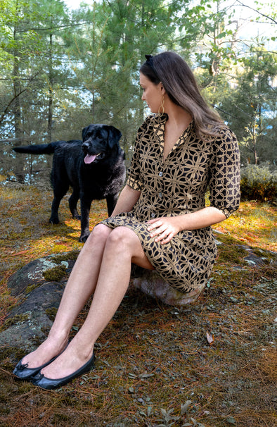 Woman in a black and brown patterned mini dress, sitting on a rock outside with a black dog in the background