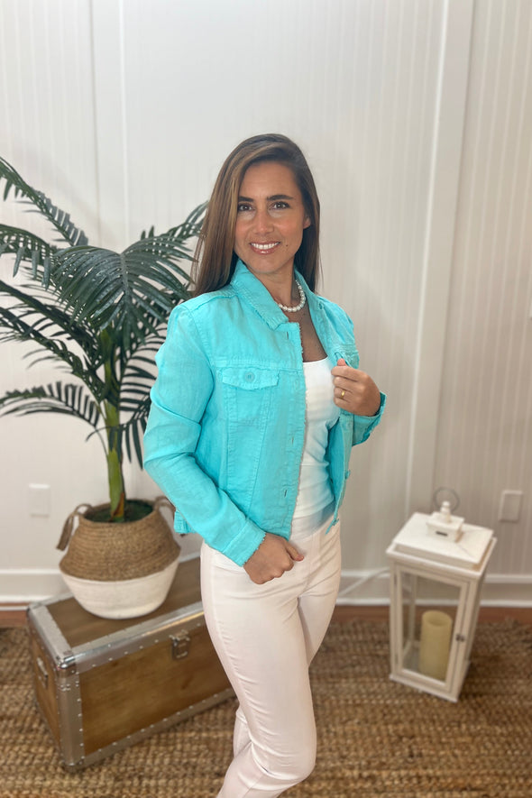 A woman stands wearing a bright aqua linen jacket with a pearl necklace, white tank top and white pants with plant and antique trunk in the background