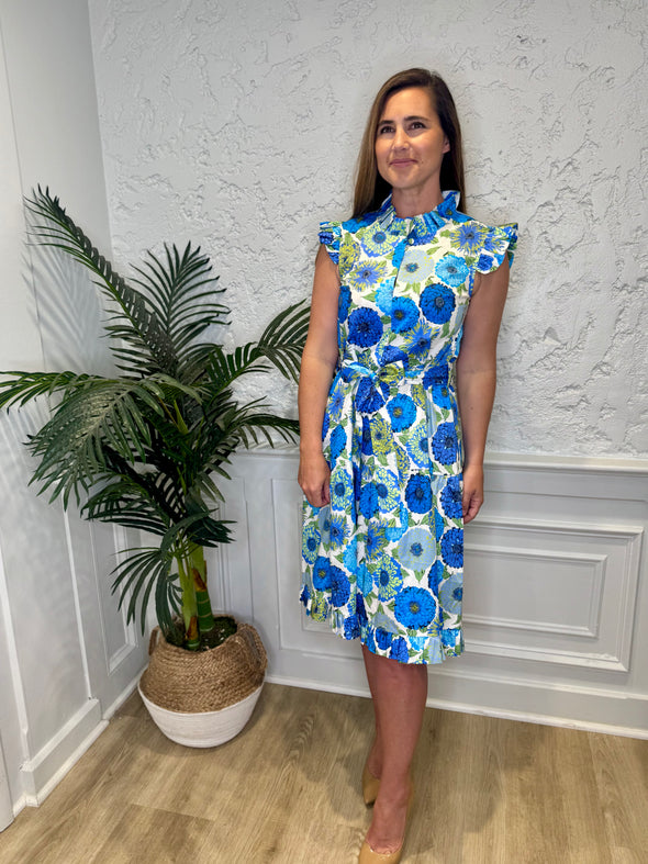 Smiling model in front of a white wall with a plant in a corner wearing the Patty Kim Megan Dress - Blue Zinnias. A midi length flutter sleeve and collar, with a tie waist and a ruffle at the bottom hem, with blue and green flower pattern