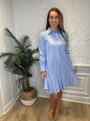 Smiling model in front of a white wall and a tropical plant in the corner wearing the Duffield Lane Iris Drop Waist Shirt Dress - Light Blue Double Stripe. A mini/knee length, long sleeve, seersucker dress. It has a collar and buttons down the front and a ruffle at the bottom hem. 